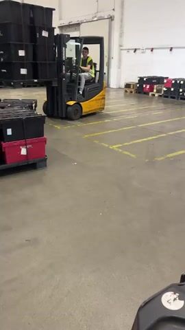 The video captures an indoor view of a warehouse environment with a yellow forklift in the center of the frame. The forklift is being operated by a person wearing a green shirt and a yellow safety vest. The forklift is carrying a tall stack of black plastic bins. To the left of the forklift, there are more black and red bins stacked on pallets. The floor is concrete with yellow painted lines indicating lanes or parking spaces. In the background, more stacks of bins and shelving units are visible. The lighting appears to be artificial, typical of a warehouse. The forklift moves forward and to the right, with the bins swaying slightly. The camera appears to be mounted on another piece of equipment, possibly a pallet jack or another forklift, positioned low to the ground and facing forward.