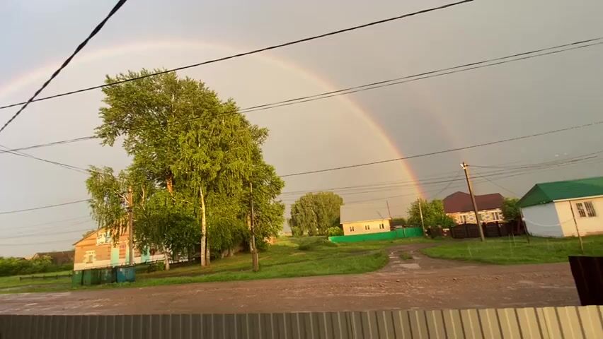 The video shows a rural scene with a double rainbow arcing across a cloudy sky. In the foreground, a dirt road is visible, wet from rain, with a corrugated metal fence in the very bottom. Behind the road, there is a green grassy area with a few trees. To the left, a two-story brick house with a blue trim is visible, along with some trash bins. To the right of the house, another house with a brown roof is seen. Further in the distance, more houses and trees are scattered across the landscape. Power lines run across the top of the frame. The lighting suggests it is either early morning or late afternoon, with the sun breaking through after a storm. There are no people or animals visible in the video.