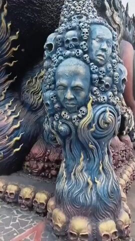 THE VIDEO SHOWS A CLOSE-UP OF A SCULPTURE. THE SCULPTURE IS DARK COLORED WITH GOLD AND BLUE ACCENTS. IT IS COVERED IN SKULLS AND FACES. THE FACES ARE RANGING FROM CHILD TO ADULT. THE SCULPTURE APPEARS TO BE MADE OF CEMENT OR A SIMILAR MATERIAL. THE SCENE IS OUTDOORS, WITH A PAVED SURFACE IN THE FOREGROUND AND BACKGROUND.
