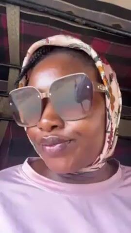 A woman with dark skin wears large sunglasses and a light pink headscarf with a floral print. She also wears a light pink shirt. She is inside a vehicle, possibly a tuk-tuk, as a red and black checkered pattern is visible in the background. The woman looks directly at the camera and slightly tilts her head. The background visible through the sunglasses appears to be an outdoor landscape.