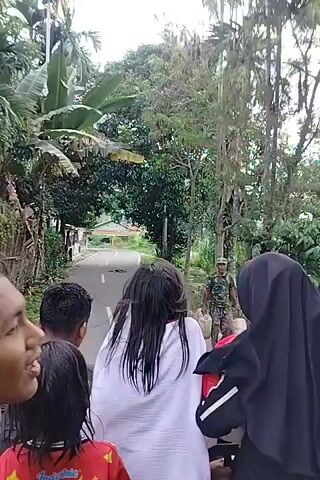 The video shows a group of people, including at least two children and two young women, on the back of a vehicle. The person in the foreground on the left appears to be a young man laughing. A young woman in a black hijab is on the right. In the center, a young woman with long, dark, wet hair is wearing a white garment. She is looking back over her shoulder. Another child with dark hair is visible. The background consists of a rural road lined with trees, including banana trees and palm trees. There are houses and vegetation visible further down the road. The overall impression is a casual scene of people traveling together.