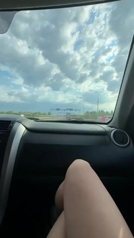 The video shows a view from inside a car driving on a highway. The dashboard is visible and a person's leg is resting on it. Through the windshield, you can see a road sign for "ИВУШКА" and "Г. КРАСНОКАМСК" with cars driving towards it, against a backdrop of a partly cloudy sky and distant greenery.  The sun appears to be out, casting shadows from the clouds.