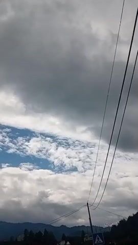 The video shows a view looking up at a cloudy sky with power lines crossing the frame. In the distance, a tree-covered hillside is visible. There are also power poles and what appear to be buildings or structures at the bottom of the frame. The camera is moving, giving a sense of travel or panning.