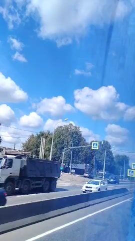 The video shows a view from inside a vehicle, looking out at a road on a sunny day. The sky is blue with scattered white clouds. On the right side of the road, there are pedestrian crossing signs. There are trees and streetlights along the side of the road. Several cars are visible on the road, including a white car on the left and a dark car in the middle. The road appears to be divided by a concrete barrier.