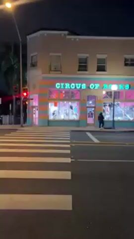The video shows a nighttime view of a street, captured from a sidewalk. The camera pans from a corner building with the sign "CIRCUS OF BOOKS" on it, across the street. The building has a colorful exterior with horizontal stripes of pink, blue, and teal. The camera moves to the right, showing a street lined with parked cars and buildings. Streetlights illuminate the scene, and strings of colorful lights hang overhead. Several people can be seen walking on the sidewalks and near the buildings. The overall atmosphere is calm and urban.