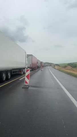 The video shows a traffic jam on a wet highway. A long line of trucks is stopped in one lane, marked off by traffic cones. A dark blue car towing a white camper passes the stopped trucks in the adjacent lane. The sky is overcast and gray. The video appears to have been taken from the side of the road. There is a grassy embankment on the right side of the road. In the distance, trees and hills are visible.