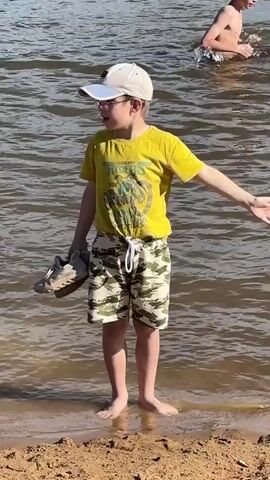 A young boy wearing a yellow t-shirt, camouflage shorts, and a white baseball cap stands at the edge of a body of water. He is holding a pair of light-colored shoes in his left hand. The boy is barefoot and his feet are in the shallow water at the edge of a sandy shore. In the background, another person is partially visible in the water. The water is rippling and appears to be a lake or a large pond. The boy is looking down at the water and seems to be contemplating entering it.