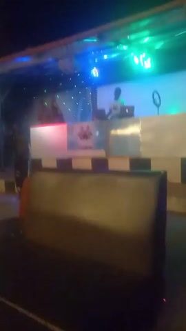 The video captures a nighttime scene at what appears to be an outdoor venue or bar with a DJ booth in the background. The DJ booth is decorated with lights and a banner that reads "SAVE HOOD." A DJ, whose face is partially visible, is seen operating equipment, likely playing music.  There are several people standing around, some of whom appear to be listening to the music or talking. The camera seems to be positioned from a seated perspective, possibly at a table with chairs and furniture visible in the foreground.  The lighting is dim, with colorful lights creating a festive atmosphere.