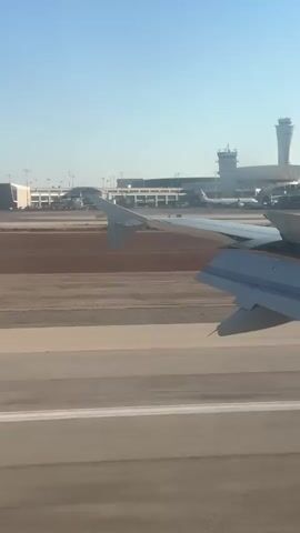This is a first-person view from an airplane window, looking out at an airport runway. In the foreground, a portion of the airplane wing is visible. Beyond the wing, a runway stretches out, marked with white lines. In the middle ground, there is a reddish-brown dirt area and a taxiway. In the background, airport buildings, including control towers, are visible, along with several parked airplanes. The sky is clear and blue. The scene suggests the airplane is either preparing for takeoff or has just landed.
