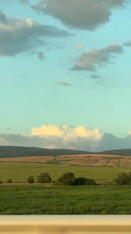 The video shows a landscape view from a moving vehicle. The scene features a vast expanse of green fields with scattered trees, leading to rolling hills and a distant mountain range under a bright blue sky with scattered clouds. The foreground shows a blurred yellow barrier, suggesting the viewpoint is from a car or train. The overall impression is one of open, rural scenery during daylight, possibly late afternoon given the soft lighting. There are no people, animals, or specific objects clearly identifiable within the frame beyond the natural environment. The video's primary focus is on the expansive natural landscape and the sensation of movement.