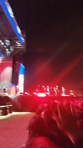 This video captures a vibrant night event taking place in Maipú, Chile. The scene is dominated by a large crowd of people gathered to witness a performance on a brightly lit stage.

The stage, located on the left side of the frame, features a metallic structure, a blue ramp or walkway leading up to it, and large screens or backdrops displaying blue and red elements. Performers, possibly a group of dancers dressed in light-colored outfits, are visible on the stage, arranged in a line or formation, moving to the music.

Intense stage lighting bathes the area in vivid colors, primarily red and blue. Powerful spotlights cut through the dark night sky, creating dramatic beams of light that are enhanced by visible smoke or haze, adding to the atmosphere.

On the right side of the frame, a dense crowd fills the foreground and extends into the mid-ground, indicating a large gathering. The crowd appears engaged, watching the spectacle unfolding on stage. The overall ambiance is energetic and lively, with loud music audible in the background, suggesting a concert or a similar entertainment event. The setting is outdoors, under the night sky, with distant structures dimly visible against the dark horizon. There is no visible text in the video.