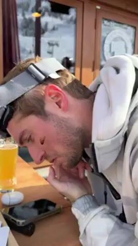 A person wearing a white jacket with a grey camouflage pattern and ski goggles pushes their finger on their face, revealing two smudges of dark color. The person appears to be sitting outdoors. Behind the person is a wooden building with a sign that reads "DOWNTOWN" with smaller text indicating "BAR MUSIC COCKTAILS." Other people can be seen in the background. The environment appears to be a sunny, mountainous area.