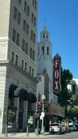 The video shows a medium shot of a street scene with a tall brick building on the left and a white tower with a cross on top on the right. The tower appears to be part of a historic theater, with a large vertical "FOX" sign in red and white letters illuminated in neon. The sign is framed with decorative colorful elements. Below the sign, there is a marquee with lights. A traffic light is visible in the foreground, showing a red light. Several cars are parked along the street. The sky is clear and blue. The overall impression is of a sunny day in an urban environment.