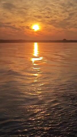 The video shows a body of water with ripples reflecting the orange sunset. The sun is low in the sky, partially obscured by clouds, casting a bright reflection on the water. The horizon is visible in the distance, with a dark silhouette of land. The scene appears to be a serene natural landscape at dusk.