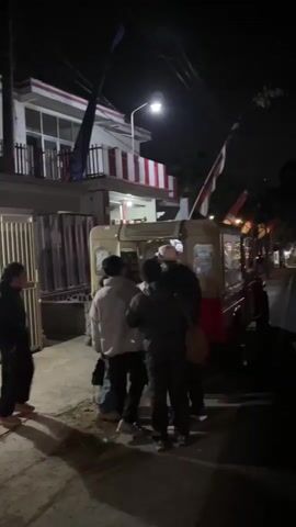 A group of six people are gathered around the back of a light-colored vehicle, possibly a jeep or utility vehicle, at night. The vehicle has a red chassis and a light-colored body with a canvas top. One person, wearing a white hat and a dark jacket, is partially inside the vehicle, seemingly interacting with the others. Three people are facing the vehicle from behind, two of whom are wearing light-colored jackets. Another person, wearing a black jacket, stands to the left of the vehicle, facing the group. One person, wearing a dark jacket, stands to the right of the vehicle, facing away from the camera. The scene is illuminated by a street light. There is a building with red and white striped trim in the background. Indonesian flags are visible, one on the building and another on a pole behind the vehicle. The ground is paved.