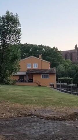 The video shows a panoramic view of a residential house and a large, ancient structure in the background, possibly a monastery or castle, nestled amongst lush green trees. The house is painted in a warm, orange hue and features a large lawn in front of it. The ancient structure, made of weathered stone, stands prominently on a hill, its silhouette visible against the pale, dawn sky. The surrounding landscape is characterized by rolling hills covered in dense foliage, with a hint of rocky terrain visible on the slope. A cobblestone path is in the foreground, leading the viewer's eye towards the scene. The time of day appears to be early morning, as indicated by the soft, diffused light and the clear sky with a gentle gradient of color. The overall atmosphere is serene and tranquil, evoking a sense of peace and timelessness. There is no visible text in the media file.