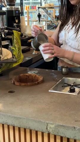 The video shows a person, likely a barista, pouring milk from a metal pitcher into a white paper cup. The person is wearing a white top, jeans, and several rings. The background includes an espresso machine, cups, and other coffee-making equipment. The counter has a unique design element – a wooden ring embedded in the surface. There is also a small silver container and a cleaning sink visible. The counter surface is stained with coffee.
