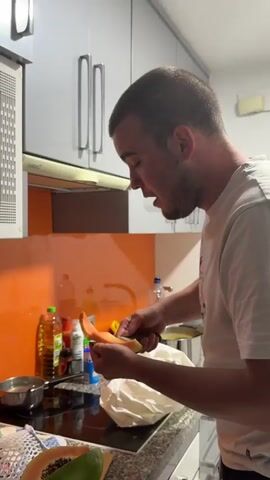 A fair-skinned man in a white t-shirt stands in a kitchen, cutting a slice from a cantaloupe half with a knife and eating it. Behind him are white cabinets, an orange wall, and various bottles and containers. A white plastic bag is on the counter in front of him. The kitchen is well-lit.