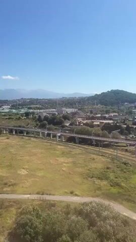 The video shows a wide aerial view of a river and its surroundings on a clear day. In the foreground, there is a dirt path and grassy areas. The river itself is muddy and appears to be flowing. On the far bank of the river, there is a stretch of green grass, followed by trees and vegetation. Further in the distance, there is a raised railway line with a train passing on it. Beyond the railway, there are buildings, industrial structures, and more vegetation. In the background, there are hills and mountains under a clear blue sky with a few scattered clouds. The camera appears to be moving, likely from a vehicle such as a train or an airplane.