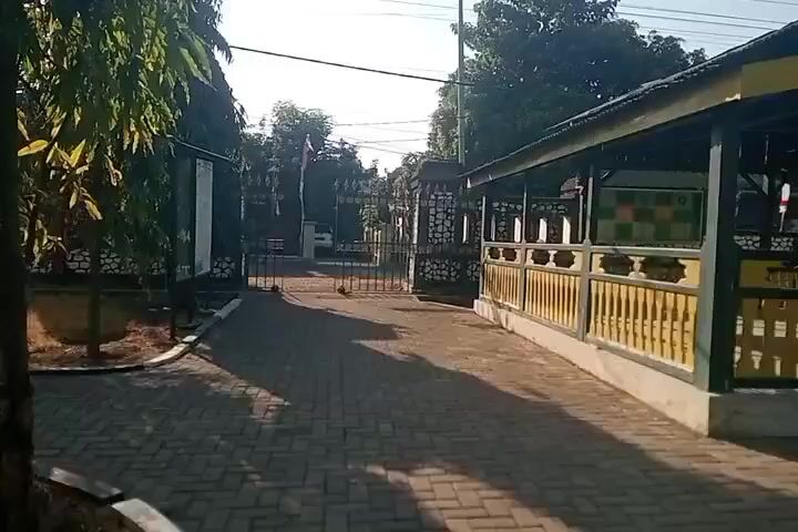 The video begins with a view of a brick-paved walkway leading towards an open gate. To the right of the walkway is a covered structure with a dark roof and yellow pillars and railings. The structure appears to be an open-air pavilion or rest area. Behind the gate, there is a stone wall and more of the pavilion. In the background, trees and utility poles are visible. The sun casts long shadows on the walkway, indicating it is likely morning or late afternoon. The scene appears to be outdoors in a well-maintained public area, possibly a historical site or a park. There are no people visible in the frame. The overall atmosphere is calm and quiet.