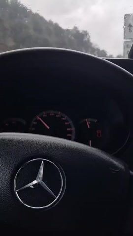 The video shows a driver's point of view while driving a car on a wet road. The car has a Mercedes-Benz steering wheel. The speedometer is visible in the lower left. The windshield wipers are on and working. The outside environment is overcast and rainy, with trees on both sides of the road. A sign indicating "El Muntanya" and "Aiguafreda" is visible overhead. Other cars are present on the road.