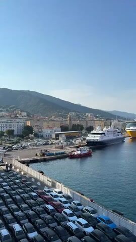 The video displays a panoramic view of a bustling port under a clear blue sky, suggesting a bright, sunny day. The foreground is dominated by a large ferry or cargo ship, its deck filled with rows of cars, indicating a significant transport operation. In the middle ground, several other vessels are docked or maneuvering in the harbor. A sleek, dark cruise ship is moored, and a bright yellow ferry with "Corsica Ferries" emblazoned on its side is also visible. A smaller red vessel, possibly a tugboat, is positioned near the dark cruise ship.

Beyond the ships, the port infrastructure is apparent, with piers, cranes, and docked vehicles. The backdrop features a picturesque coastal town nestled against rolling green hills. Buildings with terracotta roofs and white facades are visible, hinting at a Mediterranean setting. In the distance, the silhouette of a lighthouse can be seen, further reinforcing the coastal location.

The activity appears to be the loading or unloading of vehicles onto the ferry in the foreground, or perhaps the departure or arrival of various vessels in the port. The overall impression is one of active maritime commerce and travel. The clear sky and calm water suggest favorable weather conditions. The presence of multiple large ferries and a cruise ship implies a busy hub for both passengers and cargo.

Visible text includes "Corsica Ferries" on the yellow ship and "CORSICA linea.com" on the prominent red ferry. The scene is likely Bastia, France, given the visible text and the characteristic coastal landscape. The atmosphere is lively and efficient, reflecting a typical day at a major port.