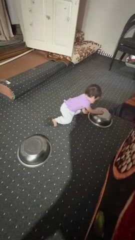 A baby wearing a purple shirt and white pants is playing on a gray carpet with a repeating pattern of white dots. There are two silver bowls on the carpet. The baby is interacting with one of the bowls, attempting to flip it over. In the background, there is a rolled-up carpet, a piece of furniture with a floral pattern, and a white door leaning against a wall. The lighting suggests an indoor environment.