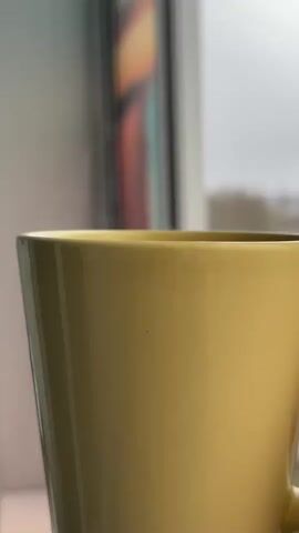 The video shows a close-up of a pale yellow mug, likely ceramic, positioned near a window. The window provides a blurred view of an overcast day with buildings and trees visible in the distance. Steam is rising from the mug, suggesting it contains a hot beverage. The background includes a partially visible item with colorful vertical stripes, possibly artwork or a decorative object. The mug is the primary focus of the shot.