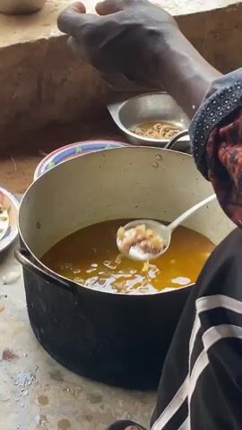 A person's hands are visible in the foreground, wearing a black and white striped sleeve and a headscarf with a red, black, and yellow pattern. The person is using a ladle to scoop food from a large, dark pot filled with a yellowish broth and pieces of meat. To the right of the large pot, there's a smaller metal bowl containing what appears to be more food. In the background, there is a rough, earthy wall and more bowls. The floor appears to be dirt or concrete. The lighting suggests it is daytime.
