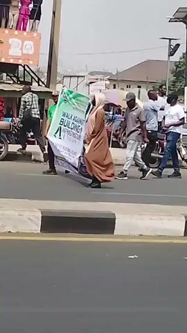 The video shows a crosswalk in what appears to be an urban environment. People are seen walking across the crosswalk, including individuals wearing traditional Muslim attire. Several auto rickshaws, also known as "Keke Napep," are visible in the background, and a white and green van is parked on the side of the road. The overall atmosphere suggests a busy, everyday scene.