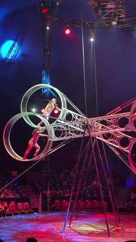A circus performance is underway under a tent. Two performers in matching tan and orange patterned costumes are on a large, rotating metal apparatus that resembles scissors. The apparatus has three large rings, two on the top and one on the bottom. One performer is standing on a platform at the top of the structure, and another performer is inside the left top ring, running to rotate it. In the bottom ring, another performer is running to rotate it. The stage floor is lit with red and blue lights. Rows of seats, mostly empty, are visible in the background.