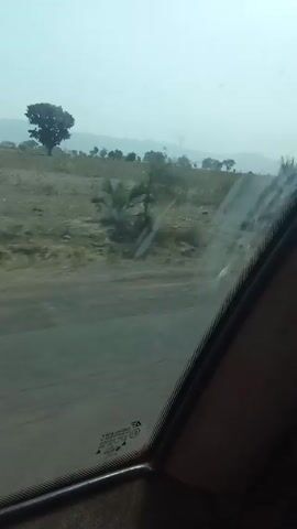 The video appears to be shot from inside a moving vehicle, looking out of the side window. The camera is focused on the passing landscape.  The scenery consists of a roadside and a grassy field with scattered trees and shrubs.  There's a haze or mist in the distance, partially obscuring what may be hills or mountains. The road surface is visible and appears to be paved, though possibly in poor condition. The window itself is somewhat dirty, with smudges visible. The interior of the car is dark, with the window frame and part of the dashboard visible. The sound appears to include music.