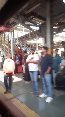 The video shows the platform of a train station with passengers. People are seen walking with luggage, sitting on benches, or waiting near the train tracks. The station has metal beams and supports overhead. Signs in Hindi and English are visible. The train is pulling into the station. The overall scene depicts the typical activity at a bustling train station.