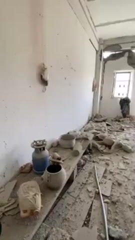 The video shows the interior of a room heavily damaged. Debris covers the floor, including what appears to be fragments of the wall and ceiling. There are shoes, plastic containers, and pieces of clothing scattered about. The walls have large holes, and the window frames are damaged. A shelf along one wall holds various objects including a blue gas canister, pots, and wrapped items.  There's also a reddish stain on the wall. The room is lit by daylight coming through the windows, indicating it's daytime.
