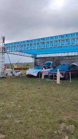 This video shows a car event taking place outdoors, likely in Antarut, Armenia, under a cloudy sky. The scene features a temporary stage structure with a blue tarp overhead, supported by metal trusses. Several cars, appearing to be electric vehicles, are displayed on the grassy ground beneath the stage. One white car has its doors open, revealing its interior.

A large crane is also a prominent feature, actively lifting a black car onto a flatbed truck. This suggests a demonstration or transport of the vehicles. The surrounding area is covered in grass, with some dirt and rocks visible. In the background, there are trees and hills, indicating a rural or semi-rural setting. The overall atmosphere seems to be that of an organized event, possibly a car show or a launch.

Visible text on a banner behind the cars reads "NIO ES8" and "N-MOTORS," identifying the brand of the vehicles. Another part of the banner contains what appears to be a website address and a phone number. The presence of the crane and the activity of moving a car suggests a dynamic and perhaps logistically complex operation. The time of day appears to be daytime, with overcast conditions.