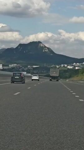 The video captures a view from a vehicle traveling on a highway. The highway has multiple lanes marked with white dashed lines. In the distance, a large mountain looms over a city with buildings scattered across the landscape. The sky is partly cloudy. Several vehicles are visible on the highway, including a large truck, a car, and a smaller vehicle. Traffic signs are present on the side of the road, indicating lane merges and prohibitions. The scene appears to be a daytime shot with clear visibility.