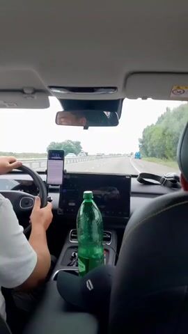 A first-person perspective video shows the interior of a car driving on a highway. The driver, wearing a white t-shirt, has their hands on the steering wheel. A smartphone is mounted to the dashboard, displaying an application. In the center console, a large green bottle of water and a black baseball cap are visible. A tablet-like screen is also mounted in the center console, displaying an image of a car. Outside the car, other vehicles can be seen on the highway, and trees line the road. The sky is overcast.