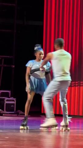 Two people on roller skates are performing on a stage. The person on the left is wearing a light blue and white skirt and top. The person on the right is wearing a white sleeveless shirt and light blue pants. They are holding hands and spinning. The background is a red curtain and black stage curtains.