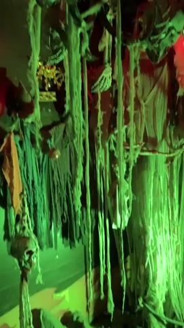 The video shows a dimly lit, green-toned room decorated with Halloween-themed props. Several fake skulls and grotesque masks hang from the ceiling by ropes and tattered fabric. In the foreground, a woman with curly brown hair laughs and claps her hands. She is wearing a black dress and sunglasses on her head. Behind her, a man wearing a baseball cap and a light-colored t-shirt smiles and holds up a small flashlight or camera, illuminating the scene. Other people are partially visible in the background. The overall atmosphere suggests a haunted attraction or a Halloween party.