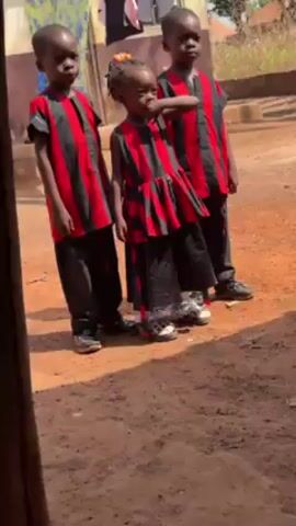 Three young children stand on a reddish-brown dirt surface. The children are dressed in matching outfits with alternating red and black vertical stripes. The two children on the sides are wearing shirts and pants, while the child in the middle wears a dress with matching pants. The child in the middle has her hand near her face, in a gesture that could be described as thoughtful or pensive. In the background, a house can be seen. The scene appears to be taking place outdoors.