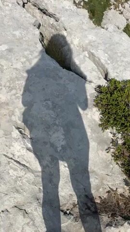 The video begins with a close-up shot of a shadow of a person standing on rocky terrain. The camera then pans up and away to reveal a mountainous landscape. The scene shows a rocky, sparsely vegetated mountainside with scattered pine trees. In the distance, a vast expanse of rolling mountains under a partly cloudy blue sky is visible. The overall impression is of a remote and rugged natural environment.