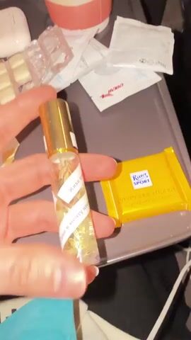 A person's hand holds a small, cylindrical bottle of perfume on a gray airplane tray table. The bottle has a gold-colored cap and contains a clear liquid with what appears to be dried flowers inside. A white label spiraling around the bottle has Russian text printed on it. Next to the perfume bottle is a small, yellow-wrapped Ritter Sport chocolate bar. In the background, the tray also holds a white container, individually wrapped pieces of gum, and several small, white packets with red Cyrillic lettering. The angle is from above, looking down at the tray.