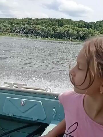 A girl in a pink shirt is in a boat. The boat is moving fast on a lake, leaving a wake behind it. The background is a shoreline with trees and a cloudy sky. The video is taken from the perspective of someone sitting in the boat.