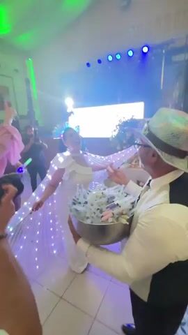 A woman in a white dress with a cape adorned with lights is being fed by a man wearing a hat and vest. The man is holding a large silver bowl filled with what appear to be rolled-up pieces of paper, possibly notes or messages, along with some small flowers or decorative items. The scene appears to be a celebration or event, indicated by the music, lighting, and other people in the background. The woman is smiling and facing the man as she accepts the item he is feeding her. The background shows a stage with DJ equipment and lighting effects.