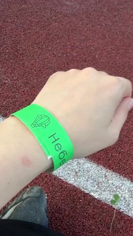 The video shows a close-up of a person's left wrist with a bright green wristband. The wristband has a black outline of a hot air balloon and the word "Небесная ярмарка" (meaning "Sky Fair" or "Heavenly Fair") written in black Cyrillic letters. The wristband is partially covering a red, granular surface that appears to be a track or sports field, marked with a white line. A dark shoe and a small green clover are visible at the bottom of the frame. The lighting suggests an outdoor setting during the day.