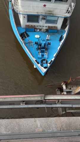 An eye-level static shot displays a top-down view of a blue and white boat moored at a pier. The boat has a prominent blue prow and a red star is visible on the upper deck. Several objects are on the deck, including a blue winch, coiled ropes, and seating. The boat's white superstructure is partially visible. The pier is made of gray concrete and metal. The water surrounding the boat is a murky brown.