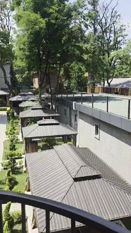 The video pans across a well-maintained outdoor area with lush greenery and several covered seating areas, likely for a restaurant or resort. A walkway is visible, lined with manicured bushes and trees. In the distance, a few people can be seen, suggesting a place of leisure. The overall impression is one of a serene and inviting outdoor space.