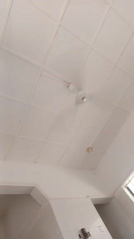 The video displays an interior room with white walls and a white paneled ceiling. A white ceiling fan with a light fixture is mounted on the ceiling, along with a second smaller, circular light. A window with a grid pattern is visible on the right side of the frame, allowing bright light to enter. A person wearing a dark cap is partially visible in the bottom right corner of the frame during parts of the video. The camera slowly pans across the upper section of the room from left to right and then back to the left.