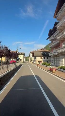 The video shows a street view from a vehicle moving forward. On the left side of the street, there is a stone wall topped with a metal fence, followed by a sidewalk and buildings. A street lamp is visible. On the right side of the street, there are multi-story buildings with balconies, some adorned with flowers. The road has a white center line and white dashed lines on the sides. The sky is blue with some wispy clouds. There are a few parked cars visible. The scene appears to be a town or village.