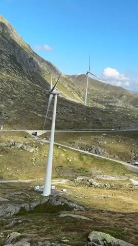 A drone video pans across a mountainous landscape with several wind turbines scattered throughout. The terrain is rocky and covered in sparse vegetation. A road winds through the valley below, and in the distance, more mountains can be seen under a clear blue sky with scattered clouds. There are no people or animals visible.